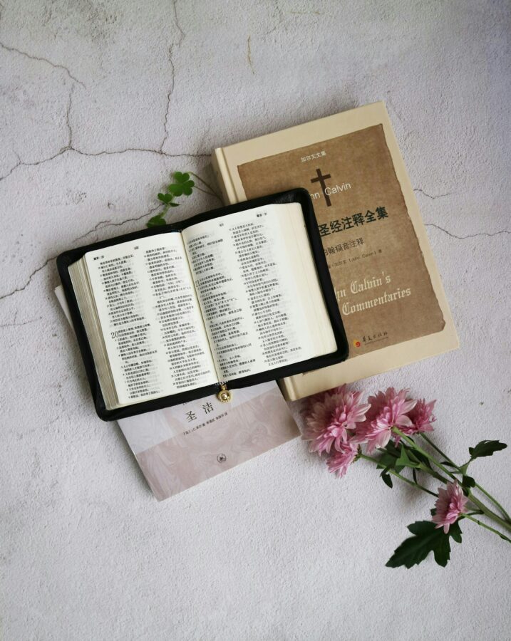 Scripture reflections Top view of an open Bible alongside books and flowers on a textured white surface.
