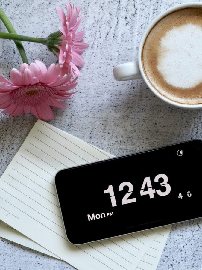 Aesthetic flat lay of a latte, pink flowers, and smartphone displaying time on a desk.