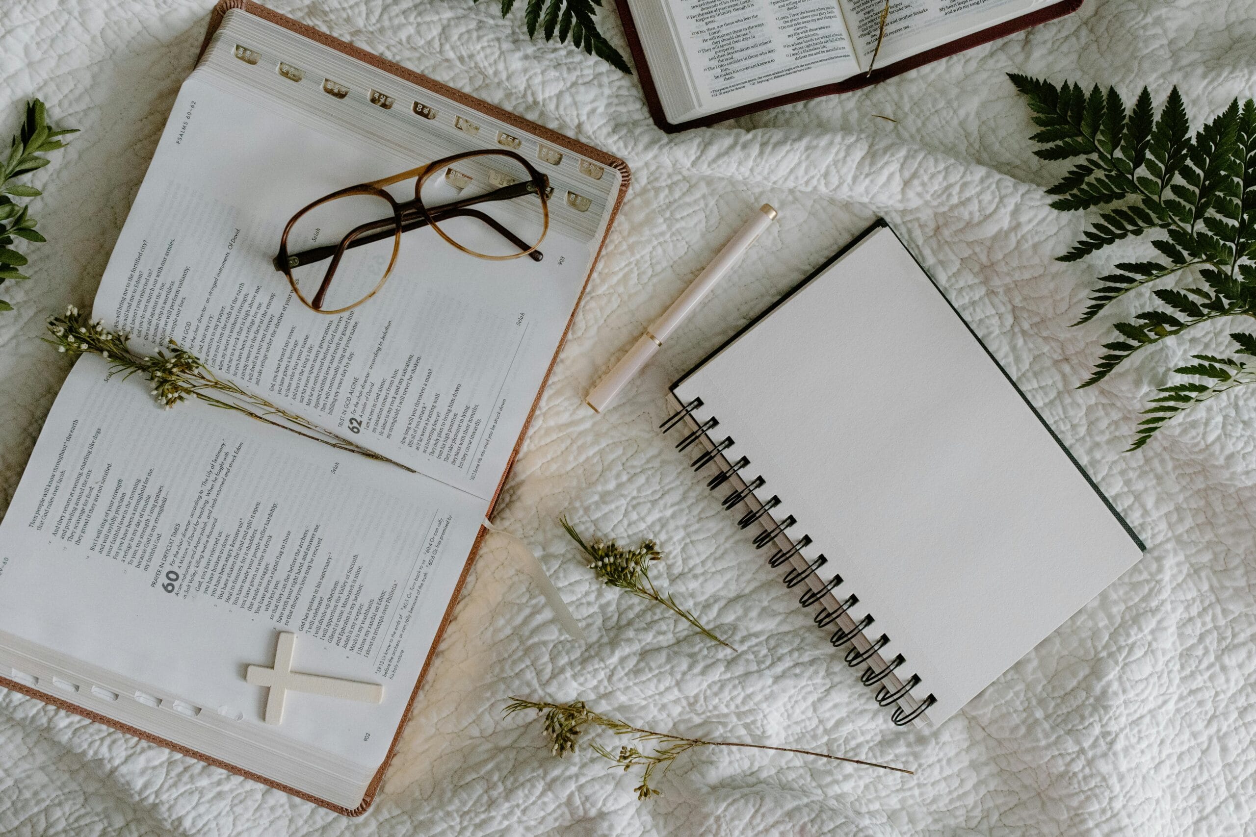 Open Bible, notebook, and eyeglasses on white bed with green leaves.
